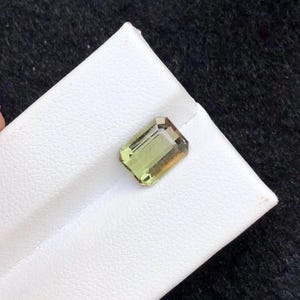 May include: A rectangular, emerald-cut gemstone featuring a gradient of light olive green to golden yellow hues. The stone has a faceted surface that reflects light, showcasing its transparent clarity and precise geometric edges.