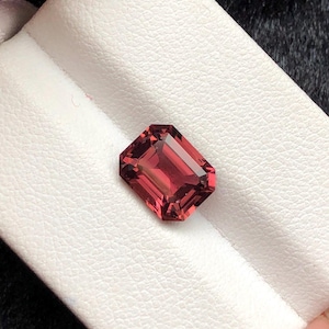 May include: A rectangular-cut, reddish-pink gemstone. The gem is set against a white, textured surface. The gemstone's facets reflect light, showcasing its clarity and color. This image highlights the beauty of the gemstone.