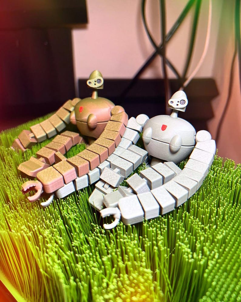 K&ouml;nnte beinhalten: Zwei kleine, bewegliche Roboter aus Plastik. Einer ist hellbraun mit goldenen Akzenten und der andere ist hellgrau. Beide Roboter sitzen auf einem Bett aus gr&uuml;nem Gras.