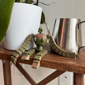 K&ouml;nnte beinhalten: Ein gr&uuml;nes und braunes, artikuliertes Roboter-Spielzeug mit einem roten Herzen auf der Brust sitzt auf einem Holztisch. Der Roboter hat lange Arme und Beine, die aus mehreren verbundenen Teilen bestehen.