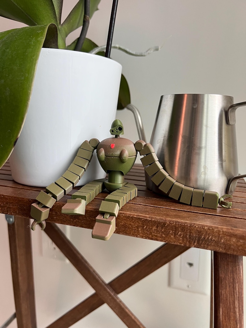 K&ouml;nnte beinhalten: Ein gr&uuml;nes und braunes, artikuliertes Roboter-Spielzeug mit einem roten Herzen auf der Brust sitzt auf einem Holztisch. Der Roboter hat lange Arme und Beine, die aus mehreren verbundenen Teilen bestehen.