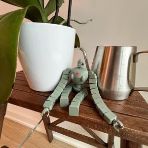 K&ouml;nnte beinhalten: Ein gr&uuml;nes Roboter-Spielzeug mit blockartigen Gliedma&szlig;en sitzt auf einem Holztisch neben einem wei&szlig;en Topf und einem silbernen Krug.