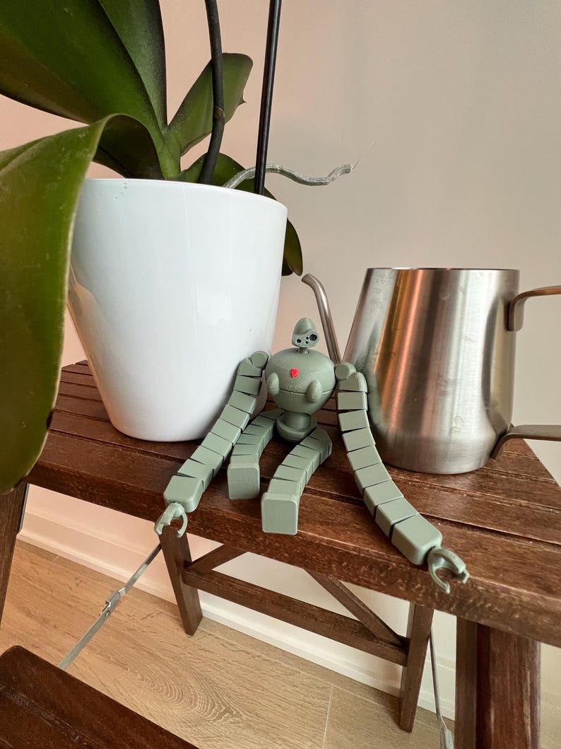 K&ouml;nnte beinhalten: Ein gr&uuml;nes Roboter-Spielzeug mit blockartigen Gliedma&szlig;en sitzt auf einem Holztisch neben einem wei&szlig;en Topf und einem silbernen Krug.