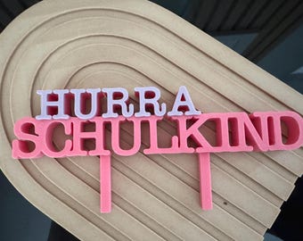 Könnte beinhalten: Ein rosa Tortenaufsatz mit den Worten "HURRA SCHULKIND" in weißen und rosa Buchstaben. Der Aufsatz ist vor einem hellbraunen, geschichteten, halbkreisförmigen Hintergrund platziert. Die Buchstaben sind erhaben und haben ein 3D-Aussehen.