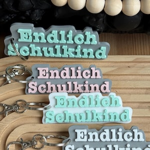 Könnte beinhalten: Vier graue Schlüsselanhänger mit dem Text "Endlich Schulkind" in verschiedenen Farben. Die Schlüsselanhänger sind übereinander gestapelt, wobei der oberste Schlüsselanhänger hellblaue Buchstaben, der zweite Schlüsselanhänger rosa Buchstaben, der dritte Schlüsselanhänger hellgrüne Buchstaben und der unterste Schlüsselanhänger weiße Buchstaben hat.