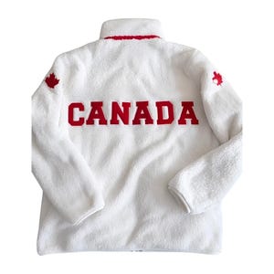 May include: A white fleece zip-up jacket featuring a high collar with red trim. The word CANADA is embroidered in large red block letters across the back. A small red maple leaf emblem is positioned on the upper portion of each long sleeve.