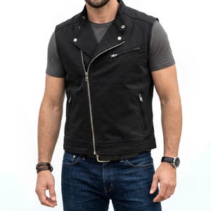 Op de afbeelding: Een zwart mouwloos vest met een asymmetrische zilverkleurige metalen ritssluiting, een opstaande kraag met drukknopen en drie zakken met rits aan de voorzijde. Het kledingstuk wordt gedragen over een grijs T-shirt en een blauwe spijkerbroek.