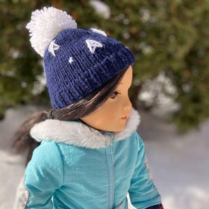 Może przedstawiać: Lalka w niebieskim, dzianym kapeluszu z białym pomponem i białymi gwiazdkami. Lalka ma na sobie niebieską kurtkę z białym futerkiem.