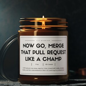 May include: A brown glass candle with a white label that reads "NOW GO, MERGE THAT PULL REQUEST LIKE A CHAMP". The candle is 9 oz and has a burn time of 45 hours.