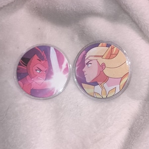 Op de afbeelding: Twee ronde, kleurrijke knopen met personages uit een tekenfilm. Eén knoop toont een rood personage met een vastberaden uitdrukking en een roze energie-explosie. De andere knoop toont een blond personage in een gouden outfit.