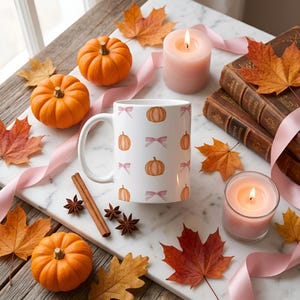 Peut inclure: Un mug en céramique blanche orné d'un motif répétitif de citrouilles orange et de nœuds rose clair. Le mug est entouré de citrouilles orange, de feuilles d'érable séchées, de deux bougies roses allumées, de cannelle, d'anis étoilé et de ruban rose.