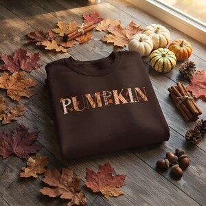 Peut inclure: Un sweat-shirt à col rond marron foncé avec le mot PUMPKIN imprimé sur la poitrine dans un motif à carreaux et feuilles aux couleurs automnales. Le vêtement est entouré de feuilles d'érable séchées, de petites citrouilles et de bâtons de cannelle.