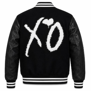 May include: A black varsity jacket featuring long black sleeves and a large white textured XO logo with a heart on the back. The jacket includes a ribbed collar, cuffs, and hem, each detailed with two horizontal white stripes against a black background.