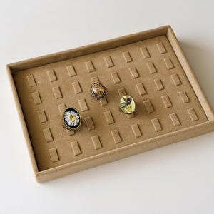 Puede incluir: Una bandeja de exhibición de joyas de terciopelo beige con ranuras rectangulares, que contiene tres anillos. Los anillos presentan diseños únicos: una margarita, un arreglo floral y un insecto amarillo y negro. La bandeja está sobre una superficie blanca.