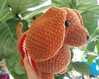 May include: A handmade brown crochet dog with black eyes and a black nose. The dog has a red and white striped band around its middle. The plush toy is soft and cuddly, perfect for children or as a decorative item.