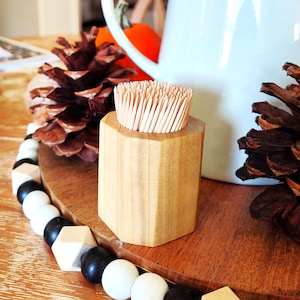 May include: A wooden toothpick holder with a hexagonal design, filled with toothpicks. The holder sits on a wooden surface, next to a pinecone and a decorative beaded garland. A white pitcher is in the background.