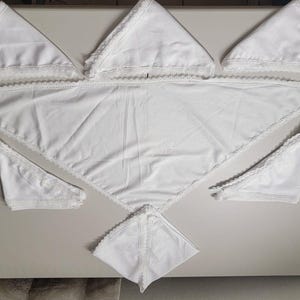 Puede incluir: Un conjunto de servilletas de tela blancas triangulares y un mantel a juego. Las servilletas y el mantel tienen un delicado ribete de encaje en los bordes. Los artículos están dispuestos sobre una superficie gris claro.