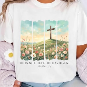 Könnte beinhalten: Weißes T-Shirt mit einem Aquarelldesign eines Holzkreuzes auf einem blühenden Hügel unter blauem und gelbem Himmel. Der Text "HE IS NOT HERE. HE HAS RISEN. Matthew 28:6" ist darunter gedruckt.