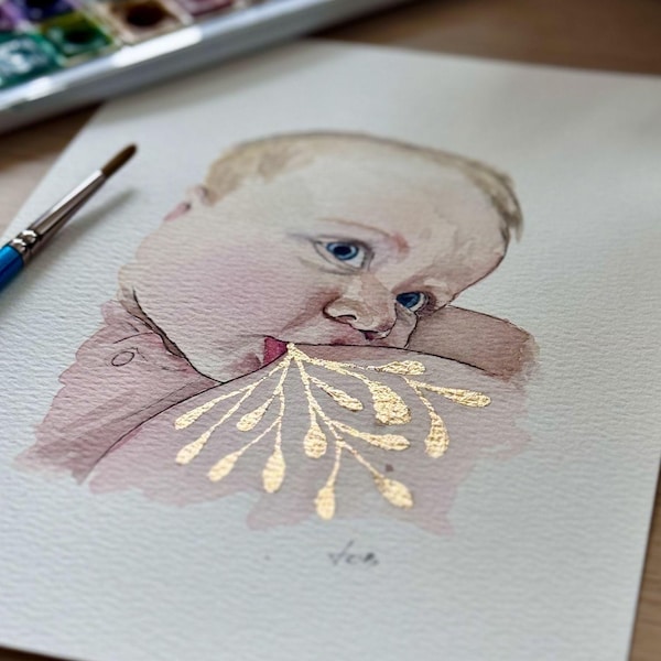 May include: Watercolour painting of a baby's face with blue eyes and blonde hair. The baby has a soft pink blush and a gold leaf design on the shoulder. A watercolour palette and brush are in the background.