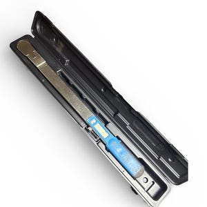 May include: A silver metal tool with a blue handle is housed in a black plastic case. The tool has a rectangular shape with a measurement scale. The case is open, revealing the tool against a white background.