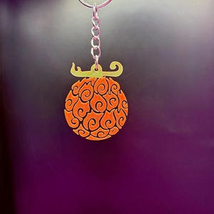 Op de afbeelding: Oranje en groene sleutelhanger met een gestileerd vlammenontwerp. De sleutelhanger is bevestigd aan een zilveren metalen ring.