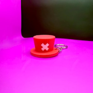 May include: A red keychain featuring a miniature hat with a white cross symbol. The hat has a circular brim and a cylindrical top. The keychain is made of a rubbery material and has a silver metal ring.