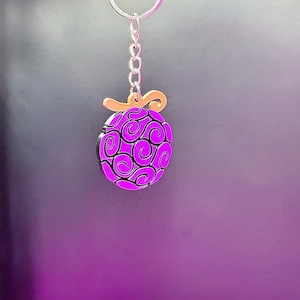 May include: A purple and gold keychain featuring a stylized design of a fruit with swirls.