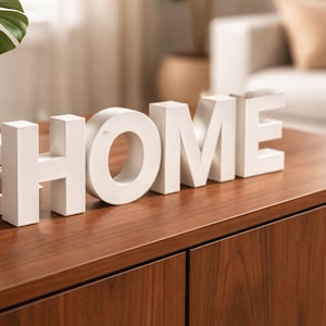 Puede incluir: Un letrero blanco con la palabra "HOME" se encuentra sobre un mueble de madera marrón oscuro. A la izquierda del letrero hay una planta en maceta con hojas verdes grandes y una pila de libros. El mueble tiene dos puertas y está frente a una pared blanca.