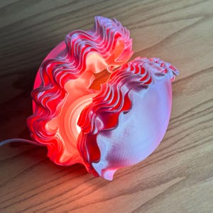 May include: A seashell-shaped lamp with a wavy, layered design. The lamp is primarily pink with red accents, and emits a warm, orange glow from within. The lamp is plugged in and sits on a wooden surface.
