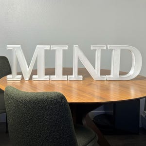 Peut inclure: Grand signe blanc en lettres majuscules épelant le mot « MIND ». Les lettres sont posées sur une table en bois ronde. Il s'agit d'un objet décoratif, idéal pour une maison ou un bureau.