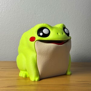 May include: A lime green frog-shaped figurine with large black and white eyes, a red cheek spot, and a beige mouth area. The frog has a smiling expression and is sitting on a wooden surface.