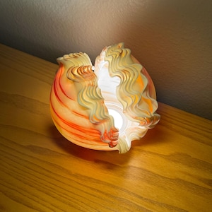 May include: A decorative glass lamp with a unique, shell-like design. The lamp features a warm color palette of orange, yellow, and white, with a wavy, textured pattern. The lamp is lit from within, creating a soft, inviting glow.
