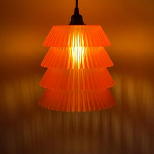 May include: Orange pendant light fixture with a tiered, pleated shade. The light fixture is suspended from a black cord and emits a warm, inviting glow. The shade's design creates a unique, layered effect, casting interesting shadows on the surrounding wall. A modern lighting solution.