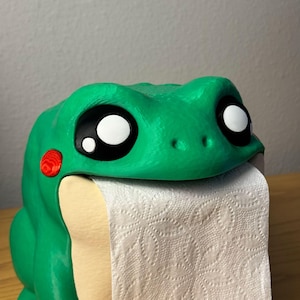 Puede incluir: Un dispensador de papel higiénico con forma de rana verde, con grandes ojos de dibujos animados y un acento rojo en un lado. El dispensador está diseñado para dispensar papel higiénico, con el rollo visible en la boca de la rana.