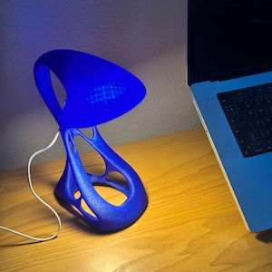May include: A blue desk lamp with an abstract design. The lamp has a curved shade and a base with open, flowing lines. A white cord extends from the base. The lamp is next to a laptop on a wooden desk.