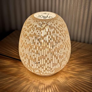 May include: A cream-colored, egg-shaped table lamp with a patterned design of vertical cutouts. The lamp is lit from within, casting a warm glow and creating a play of light and shadow on the wooden surface below. The lamp is approximately 12 inches tall.