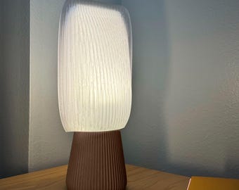 May include: A modern table lamp with a textured, off-white shade and a brown, ribbed base. The lamp is turned on, emitting a soft, warm glow. The lamp is on a wooden surface.