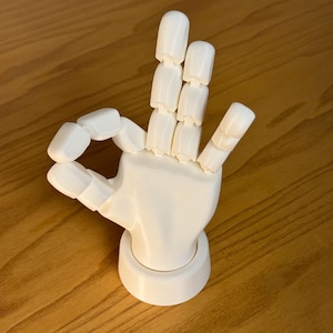 Może przedstawiać: Biały, przegubowy model dłoni w geście "OK". Palce i kciuk są lekko zgięte, a dłoń jest zamontowana na okrągłej podstawie. Model wykonany jest z gładkiego, solidnego materiału.