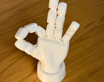 Puede incluir: Un modelo de mano blanca articulada en gesto "OK". Los dedos y el pulgar están ligeramente curvados, y la mano está montada sobre una base circular. El modelo está hecho de un material liso y sólido.