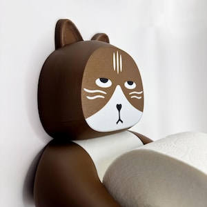 May include: A brown and white cat-shaped toilet paper holder. The holder has a cartoonish design with a grumpy expression. The holder is holding a roll of white toilet paper. The holder is mounted on a white wall.