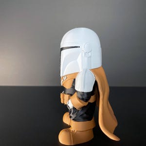 Cute, stylized Mandalorian figure in white and gold armor, set against a sandy sci-fi backdrop—an ideal collectible for fans, gift-givers, or lovers of pop culture decor.