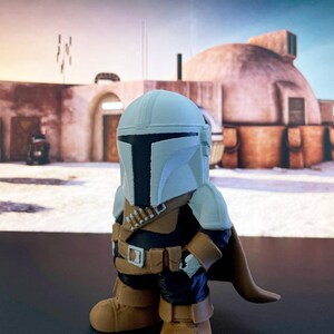 Adorable Mandalorian-inspired chibi figure posed before a Tatooine-like dome, wearing a white helmet, golden armor, and cape—ideal for Star Wars fans or geeky collectors.