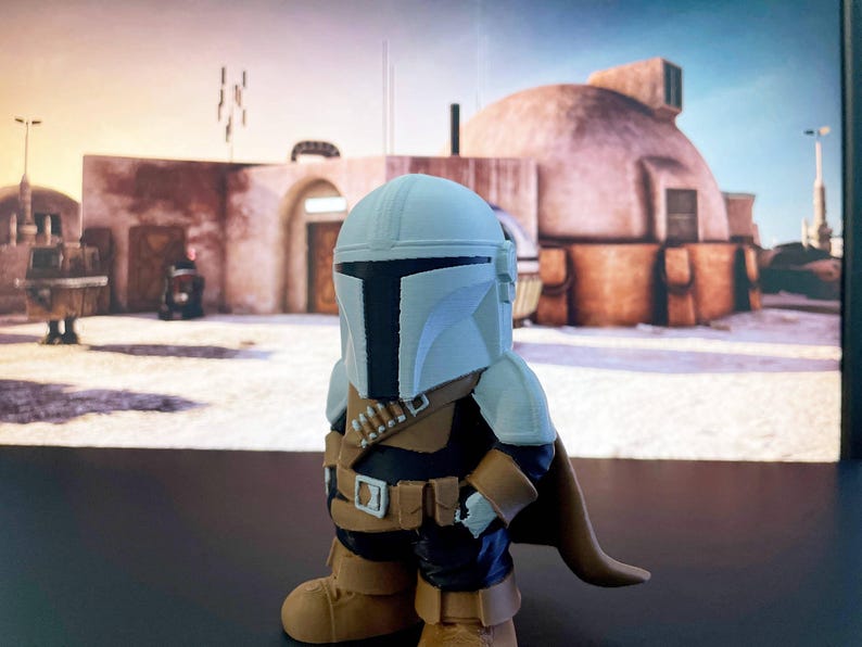 Adorable Mandalorian-inspired chibi figure posed before a Tatooine-like dome, wearing a white helmet, golden armor, and cape—ideal for Star Wars fans or geeky collectors.