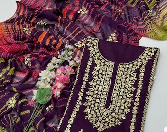May include: A dark purple salwar kameez set with gold embroidery on the top. The set includes a matching dupatta with a colorful tie-dye pattern in shades of pink, orange, and purple. A sprig of flowers is placed near the clothing.