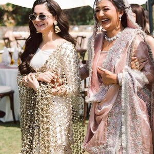 May include: Two women in elegant attire, one in a white dress with gold embellishments and the other in a pink sari with silver embroidery. They are smiling outdoors, possibly at a wedding or celebration.