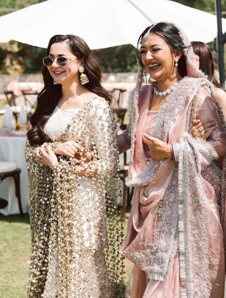 May include: Two women in elegant attire, one in a white dress with gold embellishments and the other in a pink sari with silver embroidery. They are smiling outdoors, possibly at a wedding or celebration.