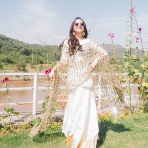 May include: A woman wearing a white dress and a gold sequined shawl, standing outdoors. The dress has a gold trim at the bottom. The background includes a lake, mountains, and flowers.