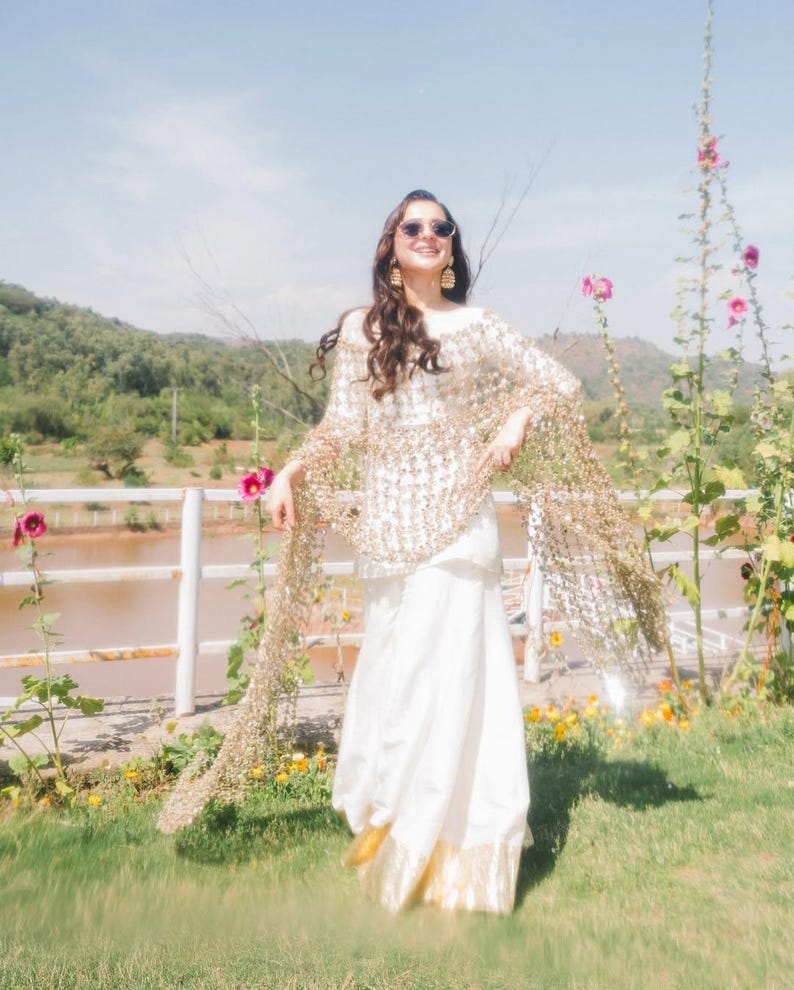 May include: A woman wearing a white dress and a gold sequined shawl, standing outdoors. The dress has a gold trim at the bottom. The background includes a lake, mountains, and flowers.