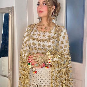 May include: A woman wearing a white and gold embroidered outfit with matching jewelry. The outfit includes a long-sleeved top and a sheer, gold-trimmed cape. The jewelry includes large earrings, a necklace, and a hand piece with a ring.
