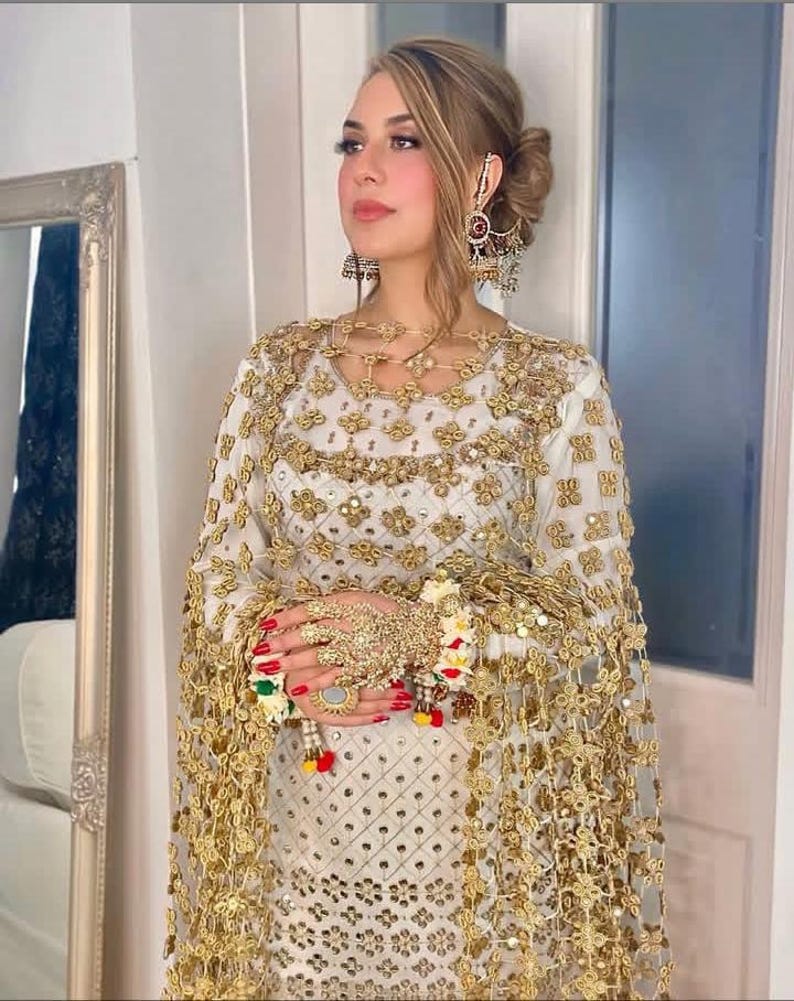 May include: A woman wearing a white and gold embroidered outfit with matching jewelry. The outfit includes a long-sleeved top and a sheer, gold-trimmed cape. The jewelry includes large earrings, a necklace, and a hand piece with a ring.